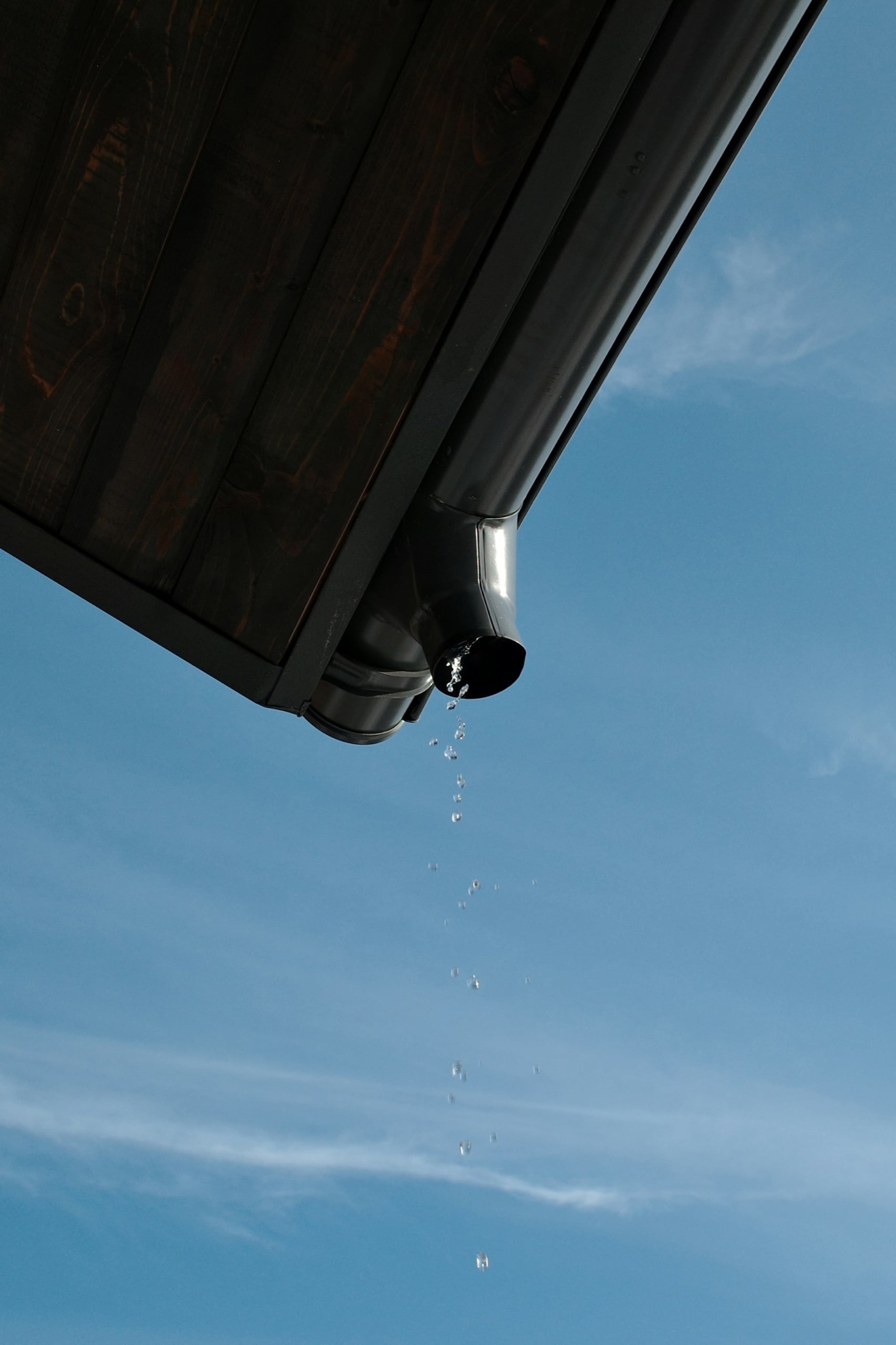 Rainwater draining from a downpipe on a home's roof gutter against a blue sky